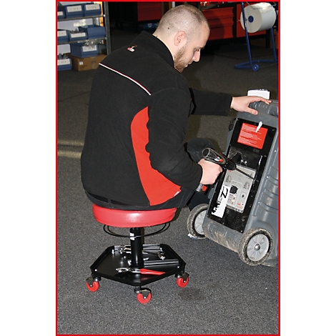 Un homme est assis sur un tabouret rouge à roulettes et travaille sur un appareil. Il porte des vêtements noirs.